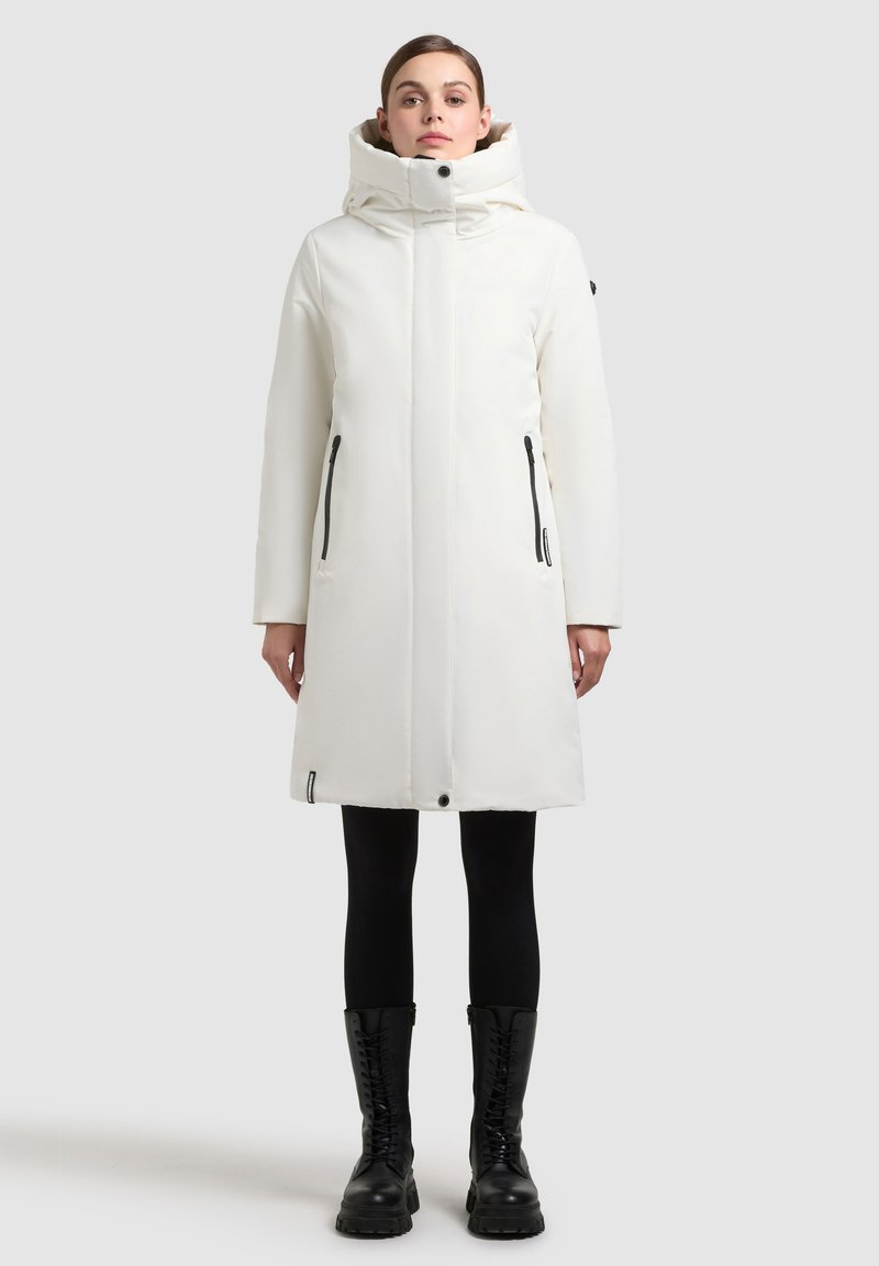 Fehér, térdközépig érő kabát magas gallérral és kapucnival. Fekete cipzáros zsebei és egyenes szabása van. Fekete leggings-szel és csizmával viselik.