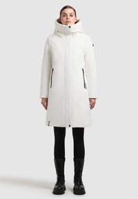 Hvid knælang frakke med høj krave og hætte. Har sorte lynlås-lommer og en lige silhuet. Bæres med sorte leggings og støvler.