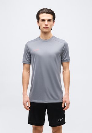 Jeune homme portant un t-shirt gris Nike et un short noir Nike, debout face à l'avant sur un fond uni.