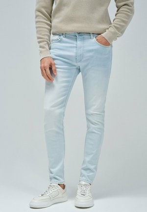 Jeans de mezclilla azul claro con un corte slim y un ligero desgaste, que incluyen bolsillos frontales y un lazo para cinturón, combinados con zapatillas blancas.