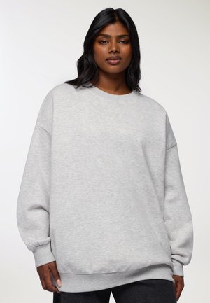 Donna che indossa una felpa grigia chiaro ampia con maniche lunghe, in piedi di fronte a uno sfondo bianco uniforme.