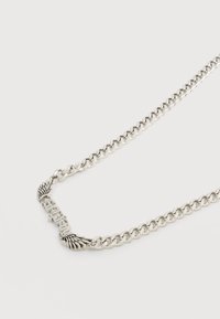 Ασημένιο κολιέ με αλυσίδα και μενταγιόν που φέρει τη λέξη "ατρόμητος", περιβαλλόμενο από χαραγμένα σχέδια φτερών.