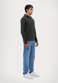 Mørkegrønn hettegenser med lommer foran og justerbar snor, parret med lyseblå rett ben jeans og hvite joggesko.