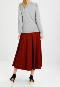 Pull gris à manches longues associé à une jupe plissée rouge longueur sol. Des chaussures à talons hauts noires complètent la tenue. Fond uni.