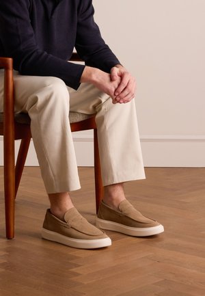 Tan slip-on shoes made of soft suede, featuring a rounded toe and rubber sole, paired with cream-colored pants on a wooden floor.