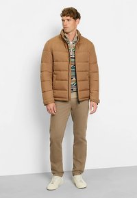 Braune Steppjacke mit horizontalen Nähten, hohem Kragen und Frontreißverschluss; darüber ein gemustertes Hemd getragen, kombiniert mit khakifarbenen Hosen und hellen Sneakern.