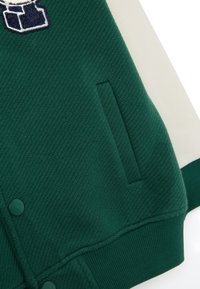 Sezione di giacca verde scuro testurizzata con una vita a coste, tasca verticale, bottoni a scatto verdi e una toppa ricamata blu navy e bianca.