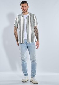 Kurzarmhemd in Weiß mit grünen Streifen, ausgestattet mit einer Knopfschließe und einer Brusttasche, kombiniert mit hellblauen Jeans und Sneakers.