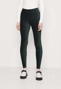 Donna che indossa leggings di velluto nero, una maglietta bianca a maniche lunghe e scarpe bianche con strappi neri, in piedi contro uno sfondo semplice.