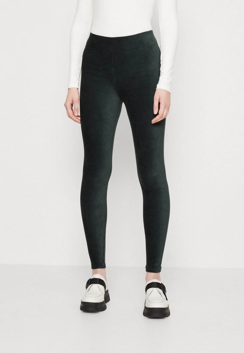 Donna che indossa leggings di velluto nero, una maglietta bianca a maniche lunghe e scarpe bianche con strappi neri, in piedi contro uno sfondo semplice.