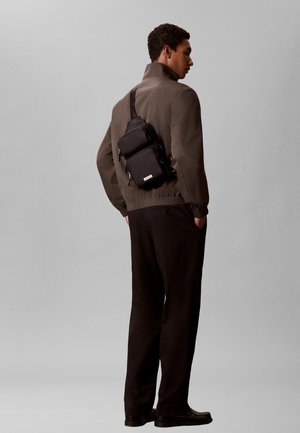 Junger Mann trägt eine braune Jacke und schwarze Hose, steht seitlich mit den Händen in den Taschen und trägt eine schwarze Umhängetasche auf dem Rücken.