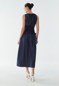 Vestido midi azul marino con cinturilla fruncida, con pliegues suaves y escote redondo. Combinado con sandalias blancas de tacón.