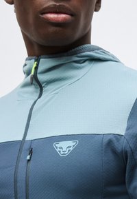 Άνδρας που φοράει ζακέτα με κουκούλα και φερμουάρ, με φωτεινό γαλάζιο πάνω μέρος, σκούρο μπλε κάτω μέρος και λογότυπο με στυλιζαρισμένο πρόσωπο ζώου στο στήθος.