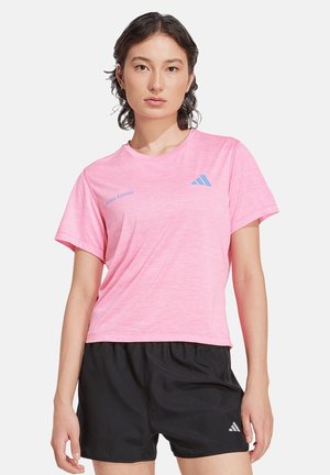 Mujer joven con cabello corto y oscuro que lleva una camiseta de running rosa de Adidas y pantalones cortos negros sobre fondo blanco.