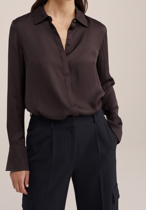 Kvinde iført mørkebrune, lange ærmer i silke med delvist åben krave, stoppet ned i højtaljede sorte bukser med lommer.