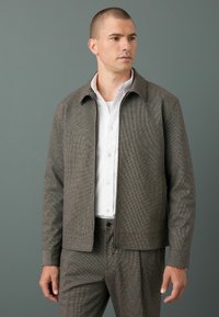 Chaqueta marrón con estampado, cuello con solapa, textura suave y cremallera frontal, llevada sobre una camisa blanca. Diseño sencillo, corte entallado.