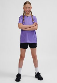 Lila atletisk t-shirt med korta ärmar, kombinerad med svarta shorts. Bärs med vita strumpor och svarta sneakers, stående mot en neutral bakgrund.