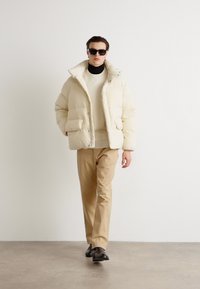 Cremefarvet pufferjakke med høj krave, forlommer, båret over en lys sweater; parret med beige bukser og sorte sko.