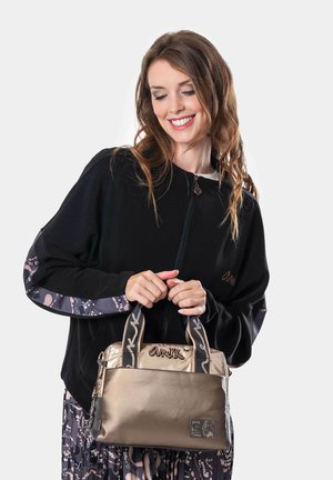 Bolso de mano de bronce metálico con tejido texturizado, dos asas y un logotipo bordado. Combinado con una blusa negra con cremallera y mangas estampadas.