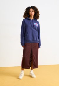 Navy hoodie with front kangaroo pocket and colorful graphic; brown skirt with side zipper and white stripes; paired with white sneakers.