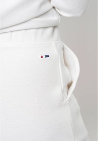 Close-up af hvide teksturerede stofbukser med et lille rødt, hvidt og blåt broderet logo nær lommen.