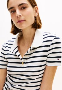 Ριγέ πόλο μπλουζάκι σε λευκό και ναυτικό μπλε, με κλασικό γιακά, τρία χρυσά κουμπιά και μικρό λογότυπο στο μανίκι. Λείο ύφασμα.