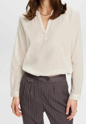 Kobieta ubrana w kremową, teksturowaną bluzkę z długim rękawem i dekoltem w serek oraz ciemnoszare, wysmuklające spodnie w prążki z wysokim stanem, dopełnione naszyjnikiem z łańcuszka.