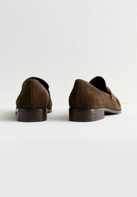 Un paio di mocassini in suede marrone con tacco basso a blocco, mostrati da dietro su una superficie chiara e uno sfondo semplice.
