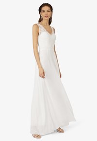 Weißes Hochzeitskleid mit Spitzenverzierungen an den Schultern, V-Ausschnitt und fließendem Rock. Glatte Stoffoberfläche mit leichter Textur. Schlichte Sandalen.