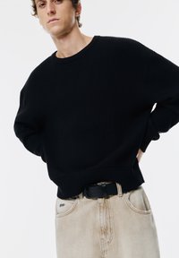 Maglione nero a coste con maniche lunghe, colletto rotondo e spacchi laterali, abbinato a jeans beige chiaro e una cintura nera.