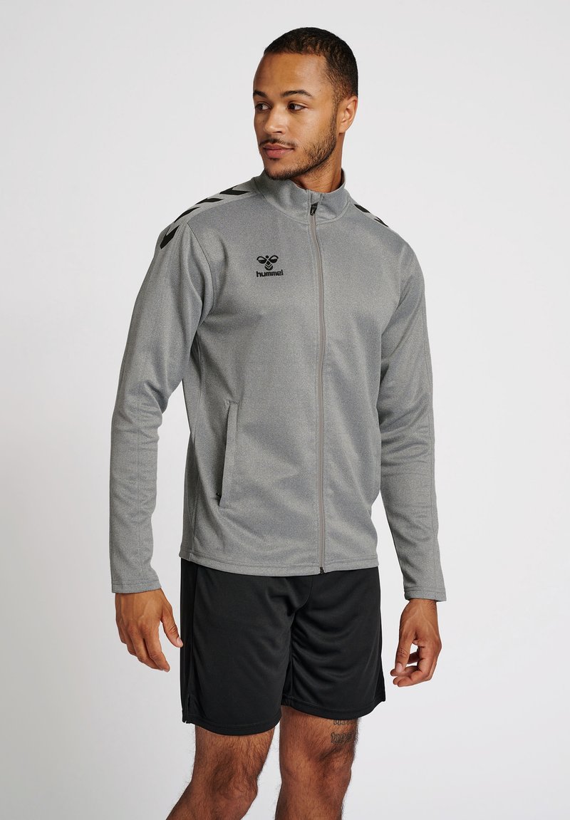 Veste de sport zippée grise avec manches longues, deux poches latérales et accents noirs. Portée par-dessus un short noir. Logo sur la poitrine.