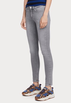 Personne portant un jean gris clair ajusté, un haut rayé bleu et noir, et des baskets bleu, marron et noir à imprimé léopard, debout sur fond blanc.