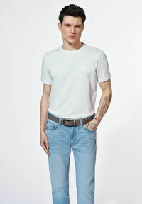 Biała koszulka z okrągłym dekoltem, krótkie rękawy, noszona z jasnoniebieskimi dżinsami i brązowym paskiem. Widoczne tatuaże na lewym przedramieniu.