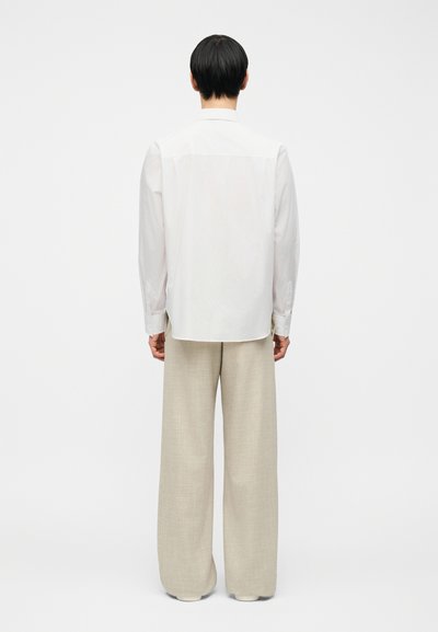 Άτομο με κοντά μαύρα μαλλιά φορούν χαλαρό λευκό πουκάμισο με μακριά μανίκια και φαρδιά μπεζ παντελόνια, στέκεται με την πλάτη γυρισμένη σε απλό φόντο.
