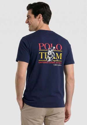 Hombre con camiseta azul marino con colorido texto "Polo Team Campeonato Español de Polo" y gráfico de jinete de caballo en la espalda, pantalones beige.