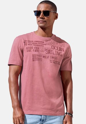 Mann mit Sonnenbrille und einem verblassten rosafarbenen T-Shirt mit nautischem Text, kombiniert mit hellblauen Jeans, vor einem einfarbigen Hintergrund.