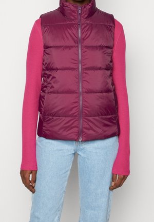 Personne portant une doudoune bordeaux zippée sur une chemise rose à manches longues et un jean bleu clair devant un fond uni.