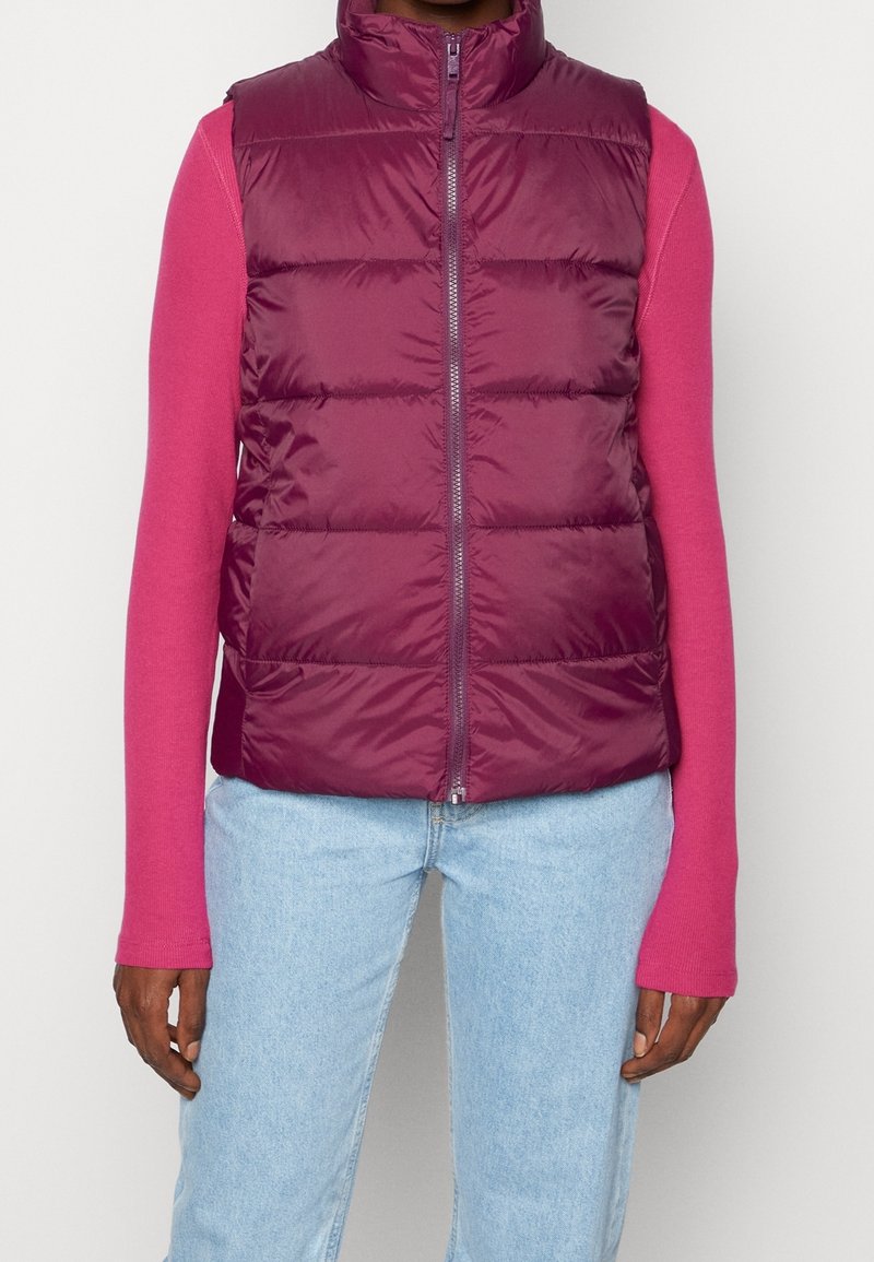 Personne portant une doudoune bordeaux zippée sur une chemise rose à manches longues et un jean bleu clair devant un fond uni.