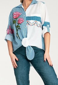 Femme portant une chemise rayée bleue avec une broderie de rose rose et des motifs bleus brodés, associée à un jean bleu, les mains reposant sur les hanches.