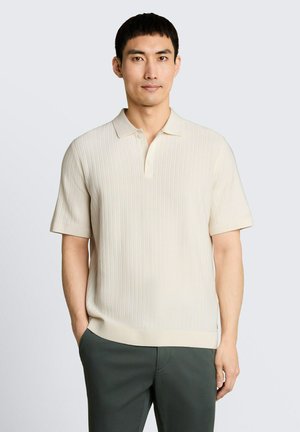 Homme portant un polo en maille crème à manches courtes et un pantalon vert foncé, debout avec une main dans la poche sur un fond uni.