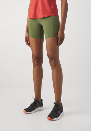 Person, die olivgrüne, figurbetonte Shorts mit seitlicher Tasche, ein rotes Sportoberteil und schwarze Laufschuhe mit lila und orangefarbenen Akzenten trägt.