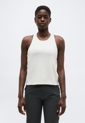 Femme debout, face à l’avant, portant un haut blanc sans manches et un pantalon noir sur un fond blanc uni.