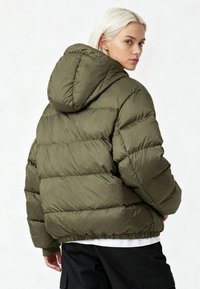 Veste matelassée vert olive avec capuche, présentant une texture matelassée, des poignets élastiques et un ourlet resserré, conçue pour la chaleur et le confort.