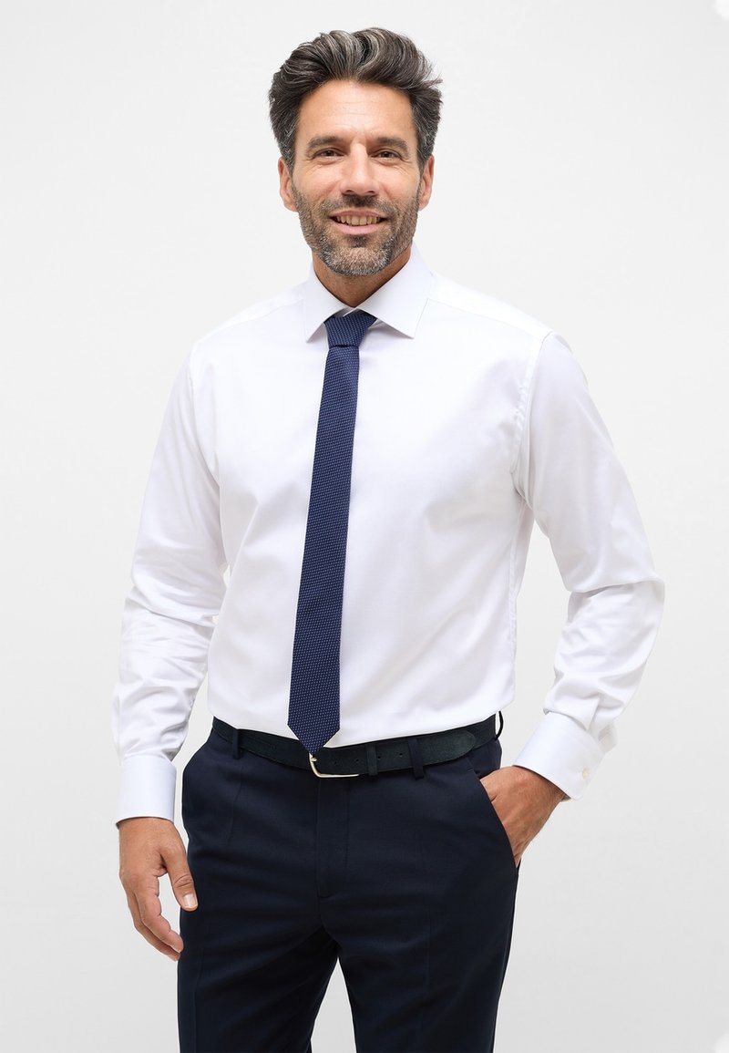 Weißes Hemd mit langen Ärmeln, kombiniert mit einer dunkelblauen strukturierten Krawatte und navyblauen Trousers. Verfügt über einen klassischen Kragen und Knopfmanschetten.
