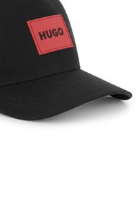 Casquette noire en tissu, avec un patch rectangulaire rouge portant "HUGO" en lettres noires en gras. Visière courbée et détails cousus.