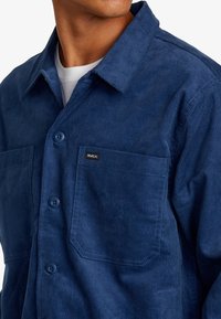 Camicia in velluto a coste blu navy con frontale a bottoni, due tasche sul petto e una piccola etichetta del marchio. Presenta un design di tessuto testurizzato e rigato.
