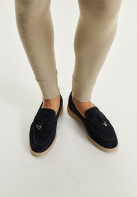Mocasinii din nubuc bleumarin cu talpă din cauciuc, având un iaht decorativ și detalii din metal auriu, asortați cu leggings bej ajustați.