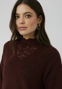Pull en maille bordeaux avec un décolleté en V profond et un détail en dentelle au col. Inclus des boucles d'oreilles circulaires dorées. Texture douce.