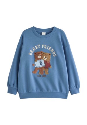 En ung dreng iført en blå "Beary Friends" sweatshirt, marineblå bukser, en hue med bjørnetema og rød-hvide sneakers står med benene spredt og armene udstrakt.