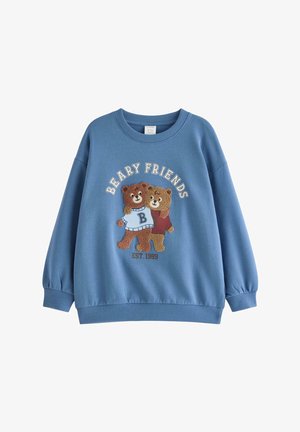 Jongen draagt een blauwe "Beary Friends" sweatshirt, marineblauwe broek, een beer-thema muts en rood-witte sneakers, staat met gespreide benen en gestrekte armen.